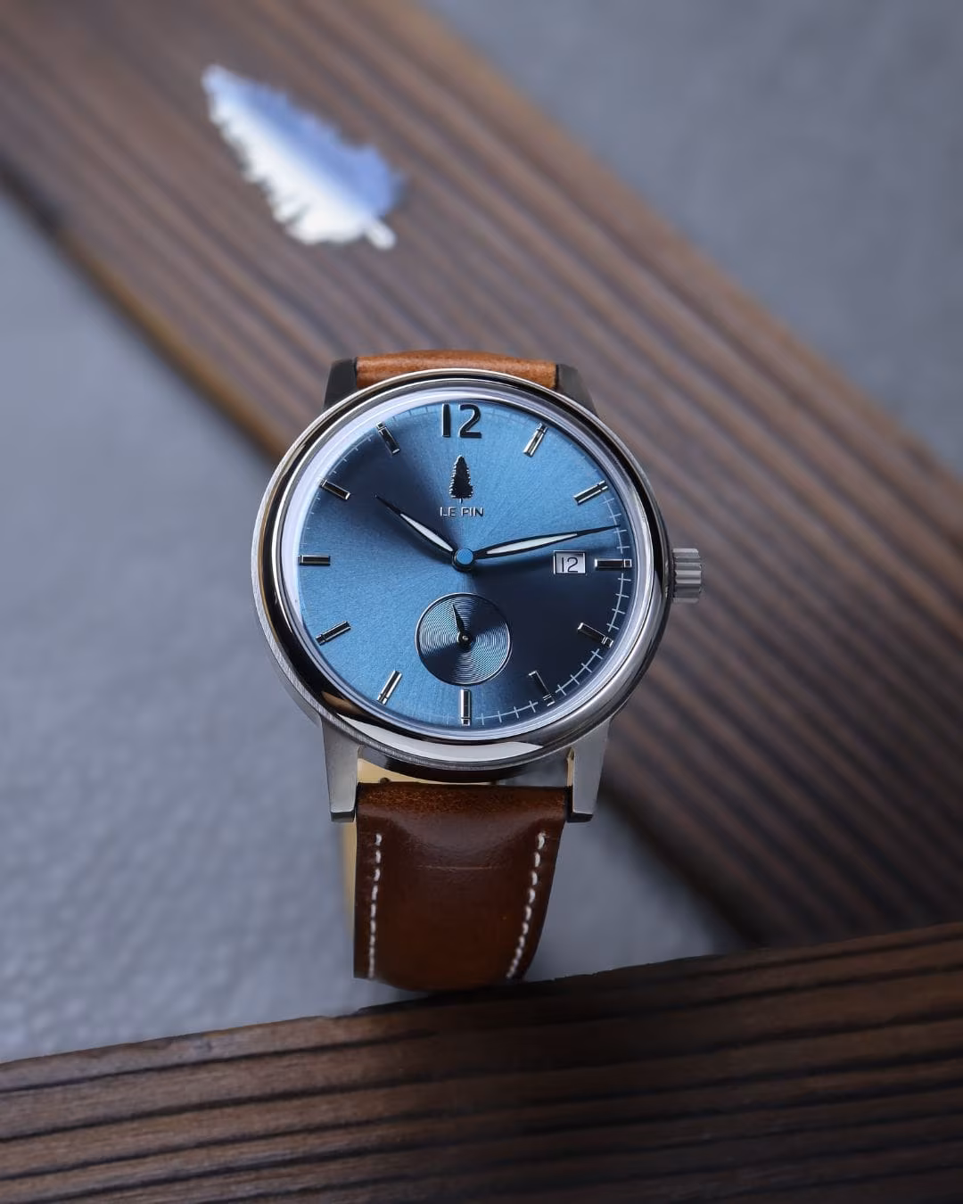 The Le Pin watch standing upright on ribbed wood beside a single pine needle.
